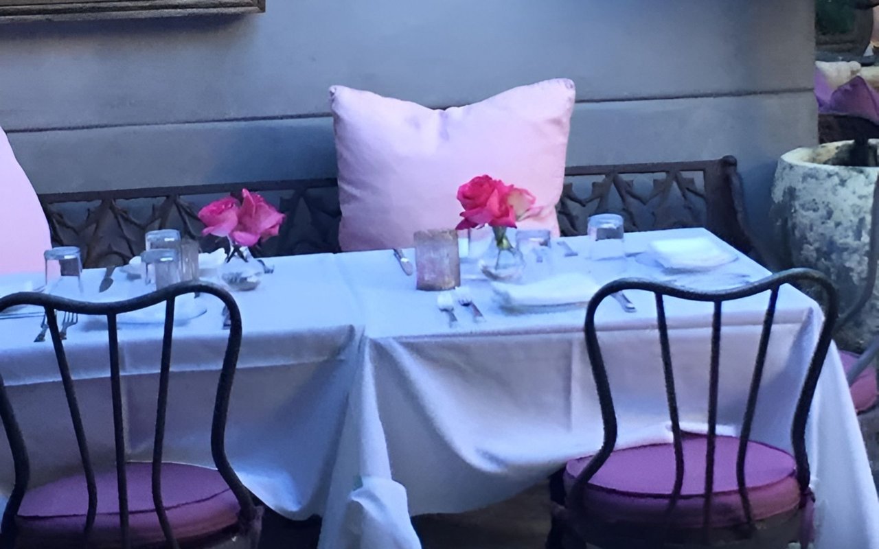 Restaurant patio table with pink flowers, a white tablecloth, and black chairs with purple cushions.