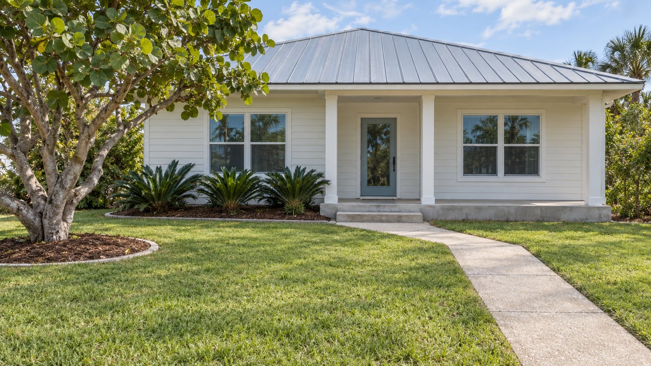 Warm white single-story cottage with a blue-gray metal roof, a raised covered entry and a deep lawn edged by sea grape and coontie planting.