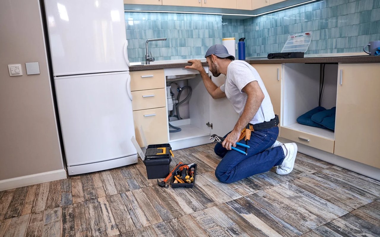 Kitchen repair worker inspecting plumbing beneath the sink, with tools on the patterned floor.