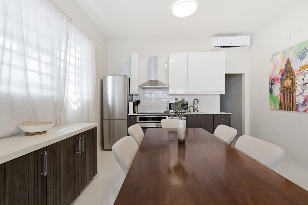 Kitchen and dining area with a long wood table, stainless steel appliances, and sheer-curtained windows.
