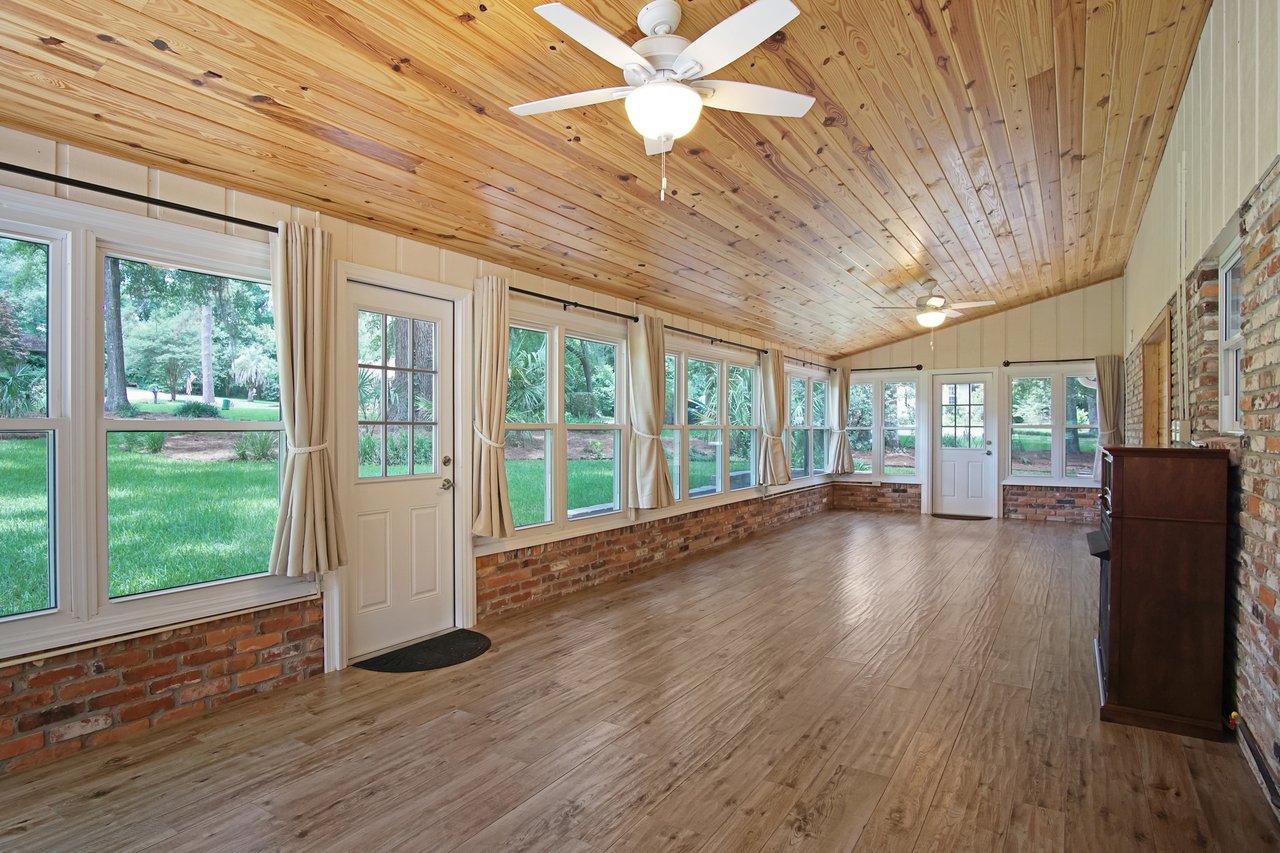 Enclosed sunroom with wood-paneled ceiling and floor, large windows with beige curtains, brick accents, ceiling fans, and views of a green lawn.