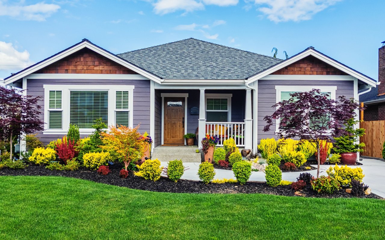 Gray single-story house with a covered porch and colorful shrubs across the front yard.