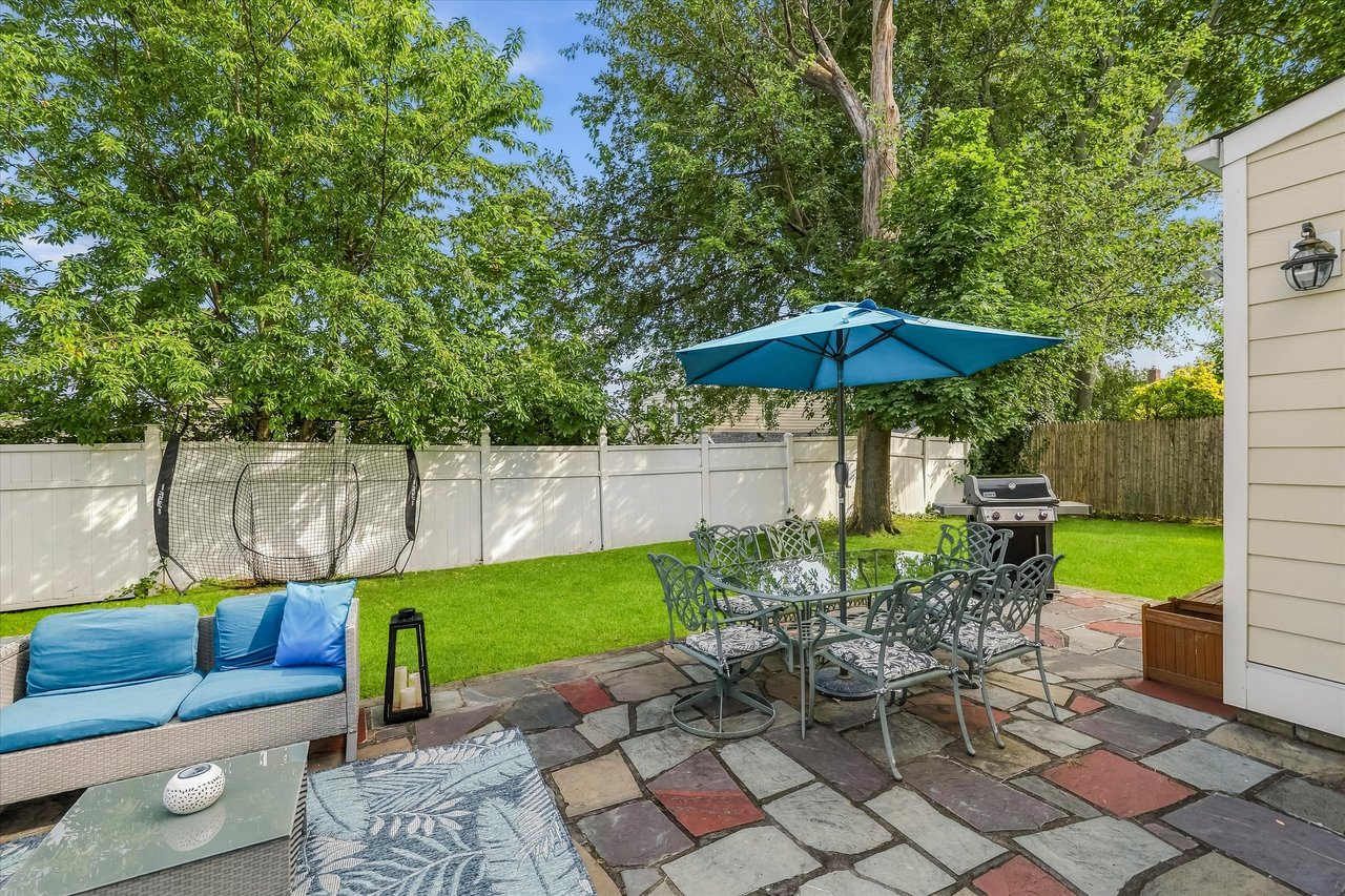 Backyard patio with stone pavers, blue seating, umbrella-shaded dining set, and fenced lawn.