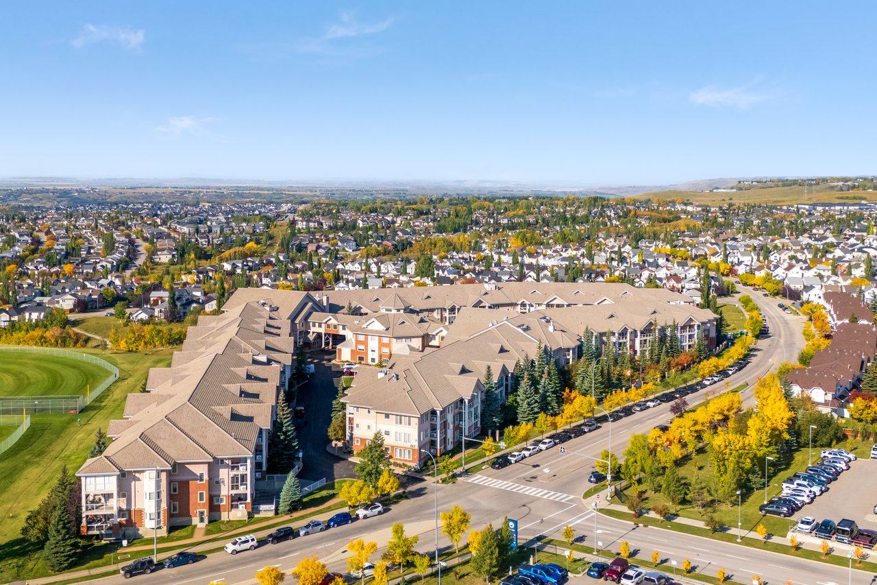 Aerial view of a multi-building condominium complex beside landscaped green space and a tree-lined road.