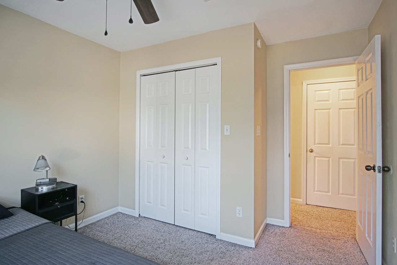 Bedroom at 529 Collinsford Road, Tallahassee, Florida, featuring beige walls, carpet flooring, and a ceiling fan. The room includes a white double-door closet, a black nightstand with a reading lamp, and a doorway leading to the hall, creating a cozy and functional layout.