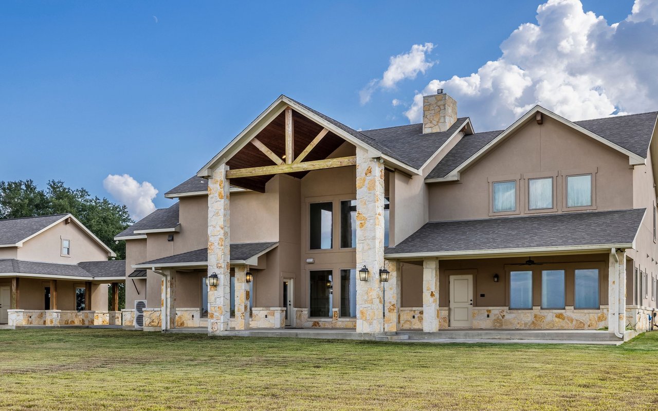 Large beige home with stone columns at the covered entry and a broad front lawn.