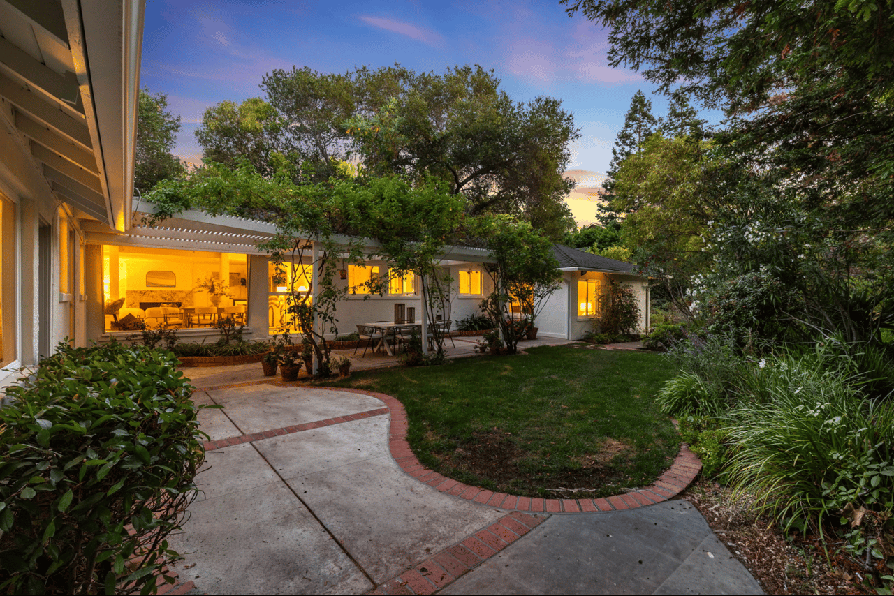 Single-story home and landscaped patio at dusk, with warmly lit windows and a curved brick-edged walkway.