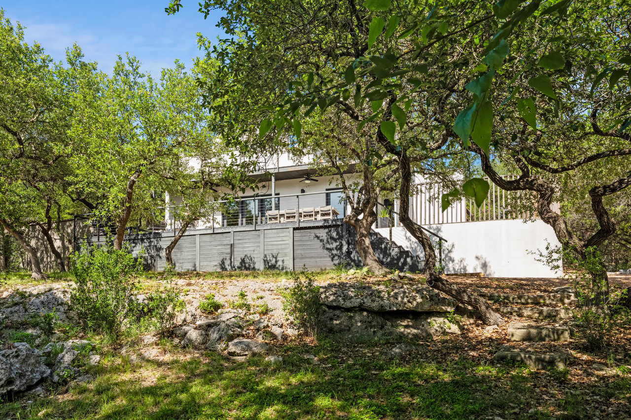Modern white home with a raised deck and outdoor dining area among leafy trees.