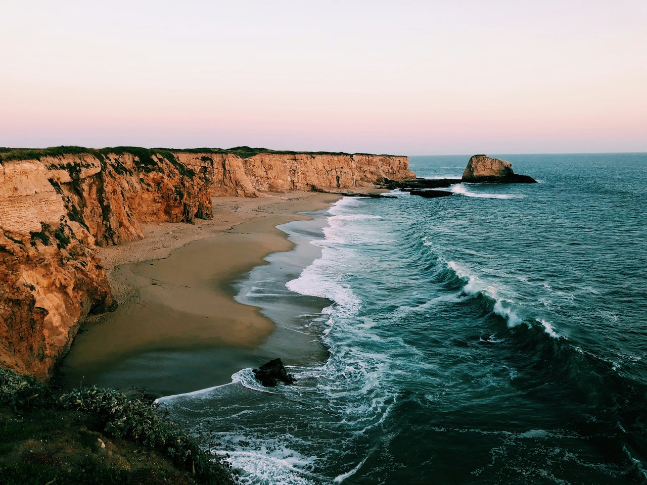 Coastal cliffs and sandy beach beside rolling waves at sunset.