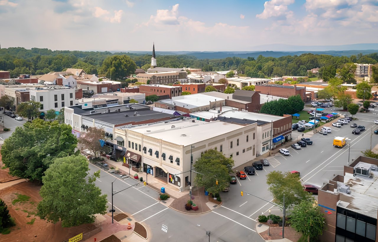 Aerial view of downtown shops and offices, with a church steeple and wooded hills beyond.