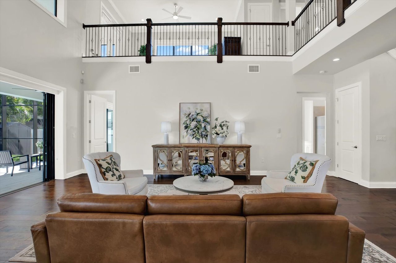 Staged living room with a brown leather sofa, white armchairs, hardwood floors, and an open upstairs gallery.
