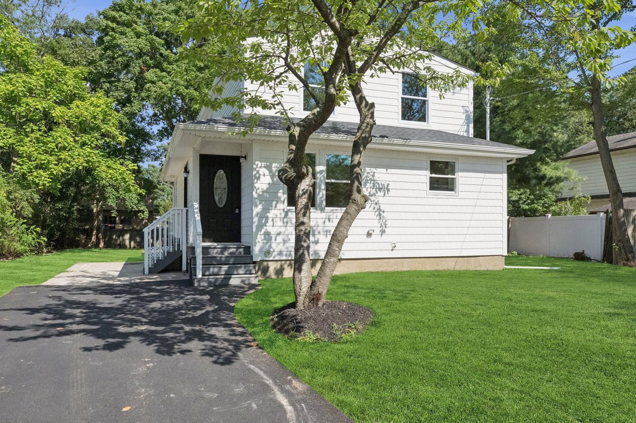 White two-story house with a black front door, driveway, lawn, and mature shade trees.
