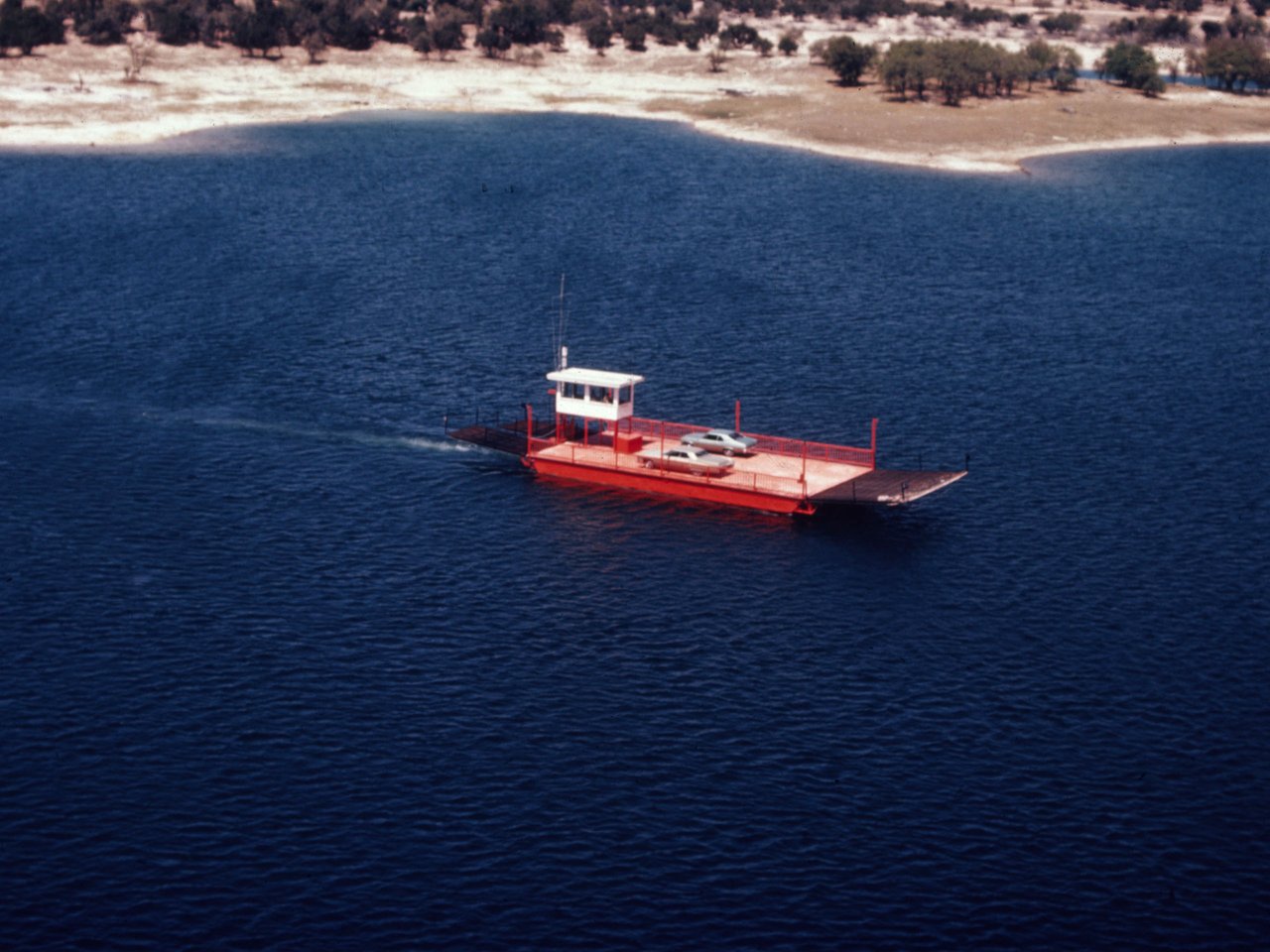 A red car ferry crosses a blue lake near a sandy, tree-lined shore.