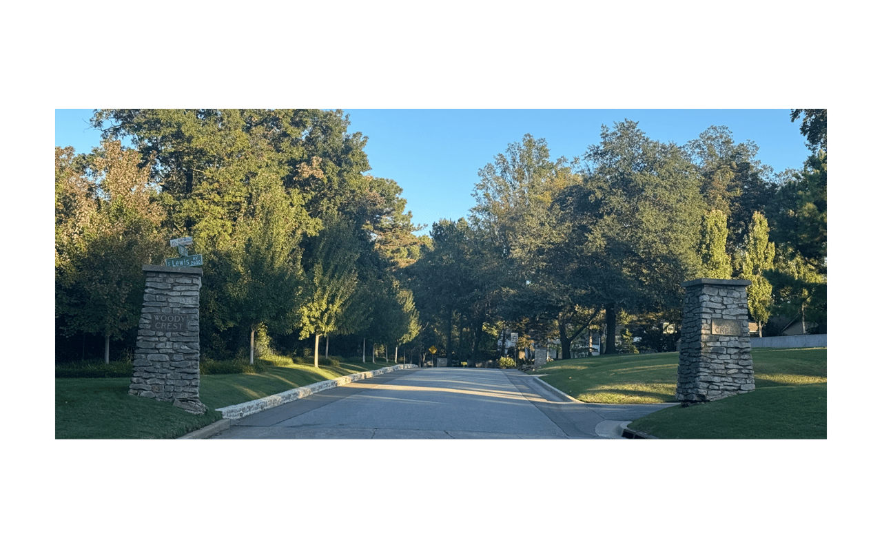 Woody Crest entrance with stone pillars and a tree-lined road in warm evening light.
