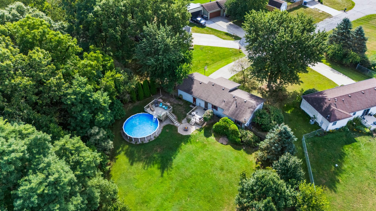 Aerial view of a single-story home with an above-ground pool on a tree-lined lawn.