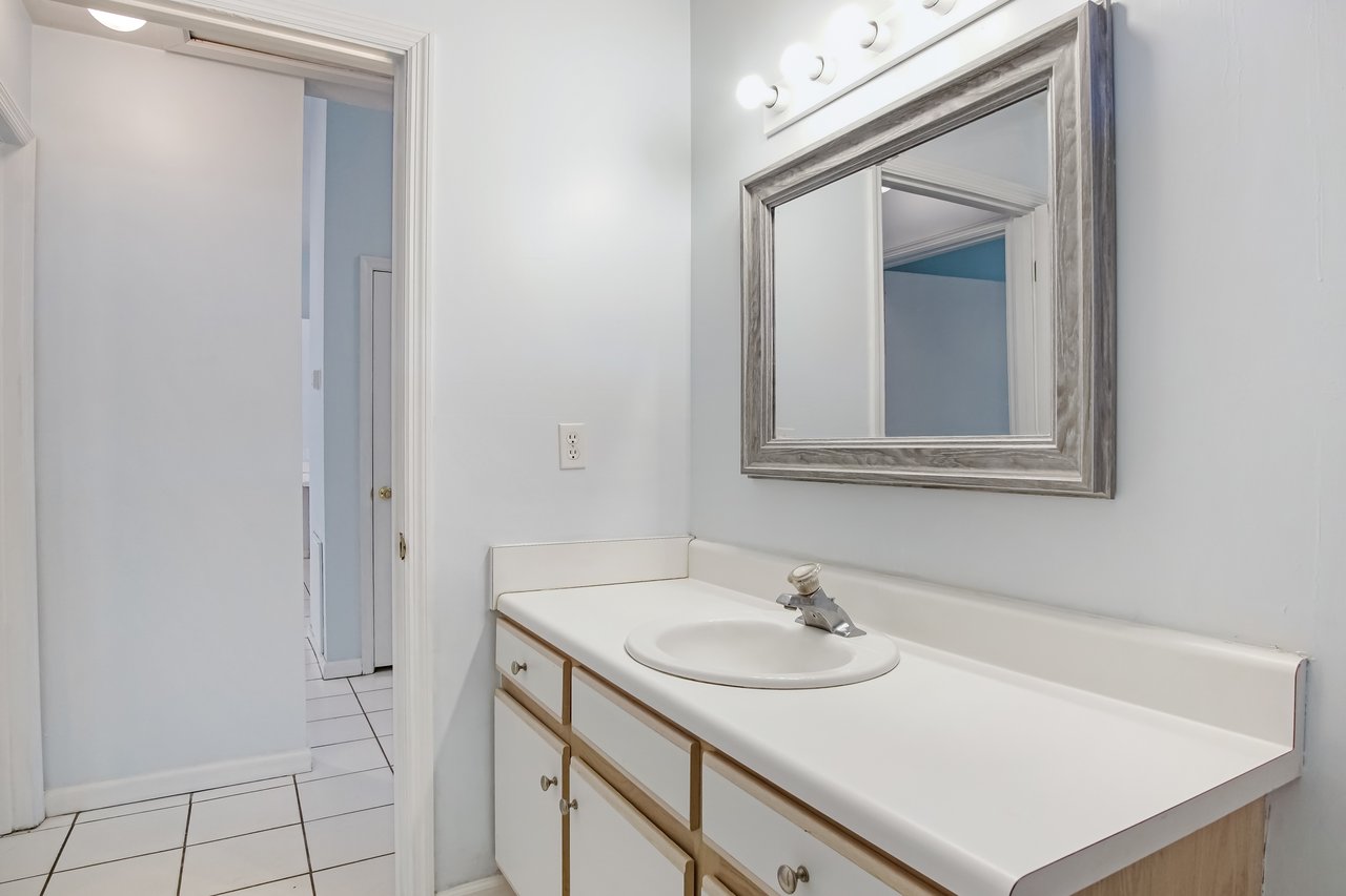 A clean, bright bathroom with a white vanity, single sink, and a rectangular mirror framed in gray wood. Overhead lights enhance the minimalist decor.