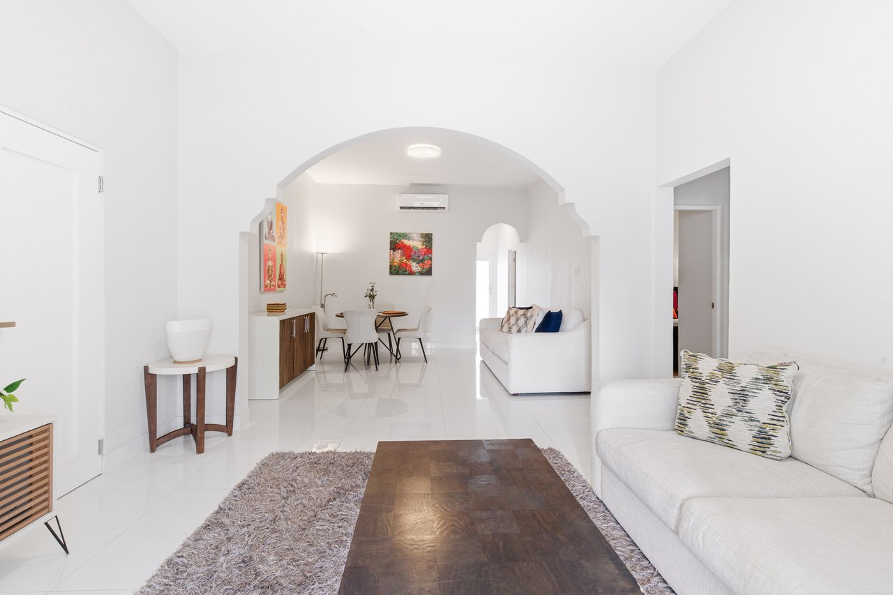 Furnished apartment living room with white sofas, dark wood coffee table, and arched opening to the dining area.