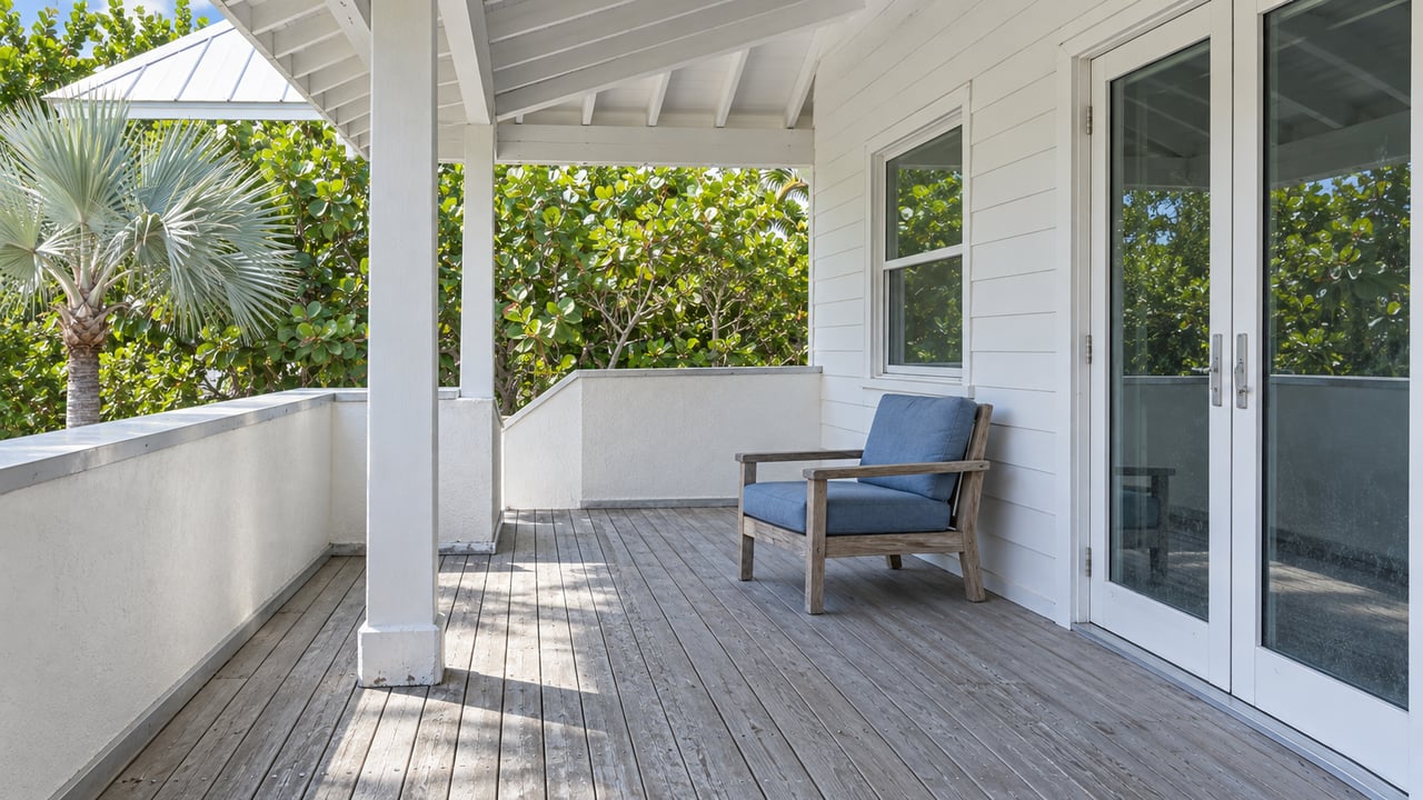 Shaded porch of a white elevated coastal home with glass doors, weathered gray decking, a teak chair and silver fan palm foliage.