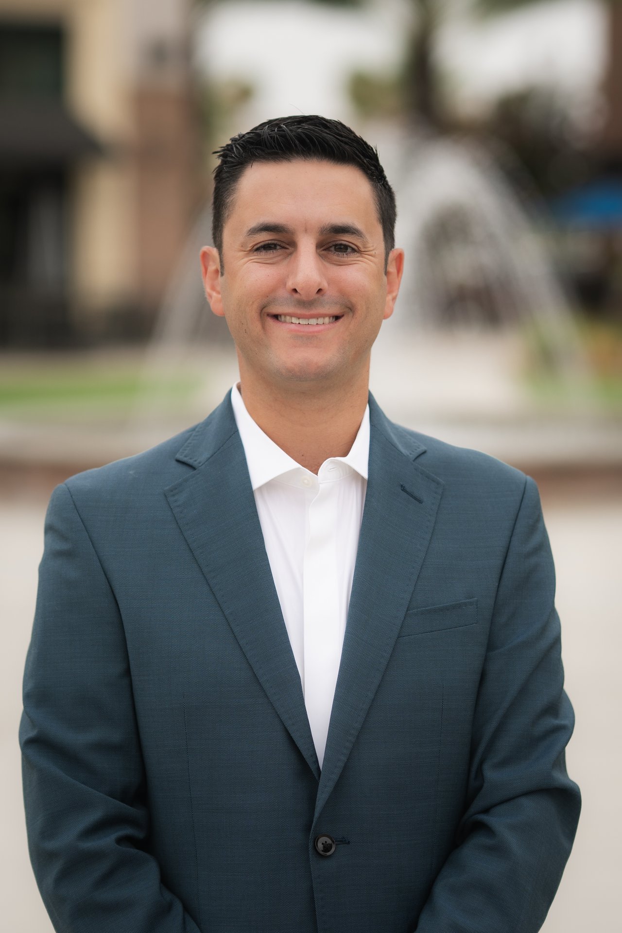 Smiling man in a blue-gray suit and white shirt, photographed outdoors.