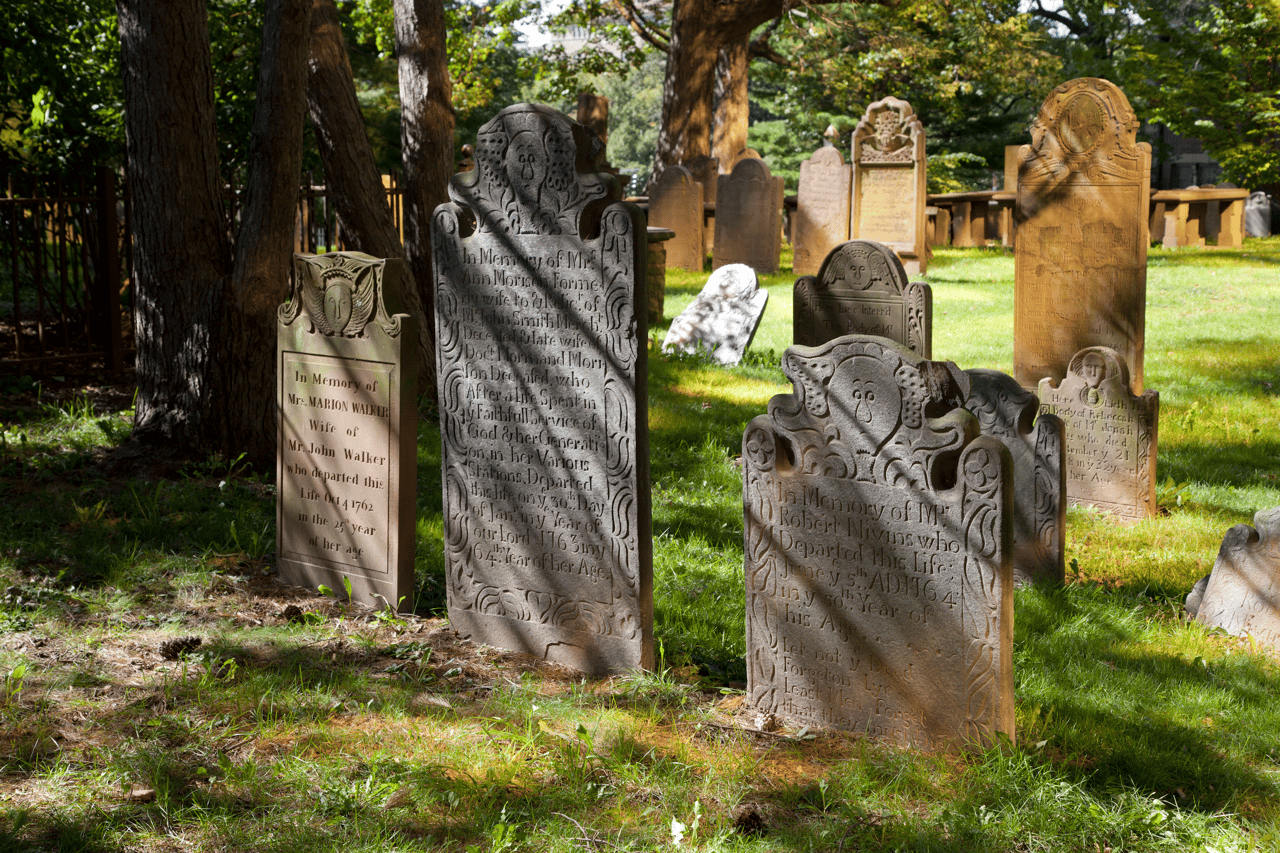 Historic carved gravestones stand among trees and grass in a sunlit cemetery.