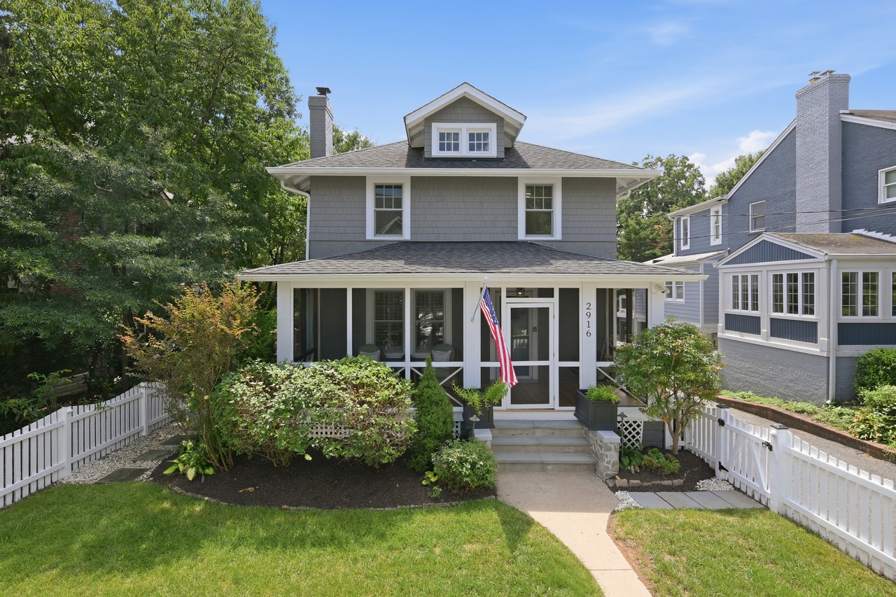 Gray two-story home with a covered porch, American flag, and white picket fence.