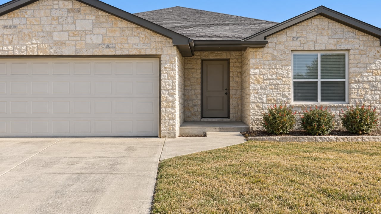 Pale limestone single-story home with a closed garage, recessed entry, concrete approach and modest flowering shrubs in clear afternoon light.