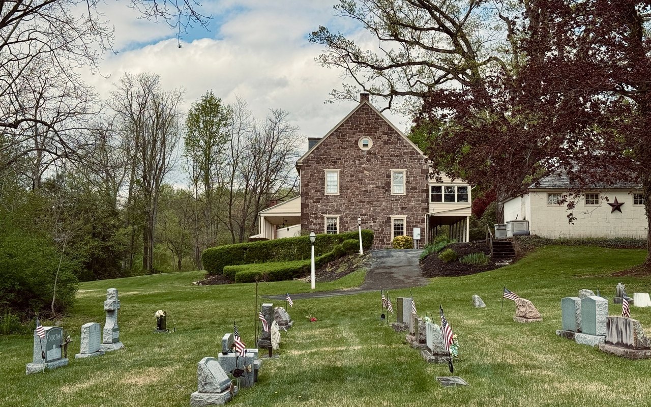Stone house beside a cemetery, with gravestones and flags across the grassy foreground.