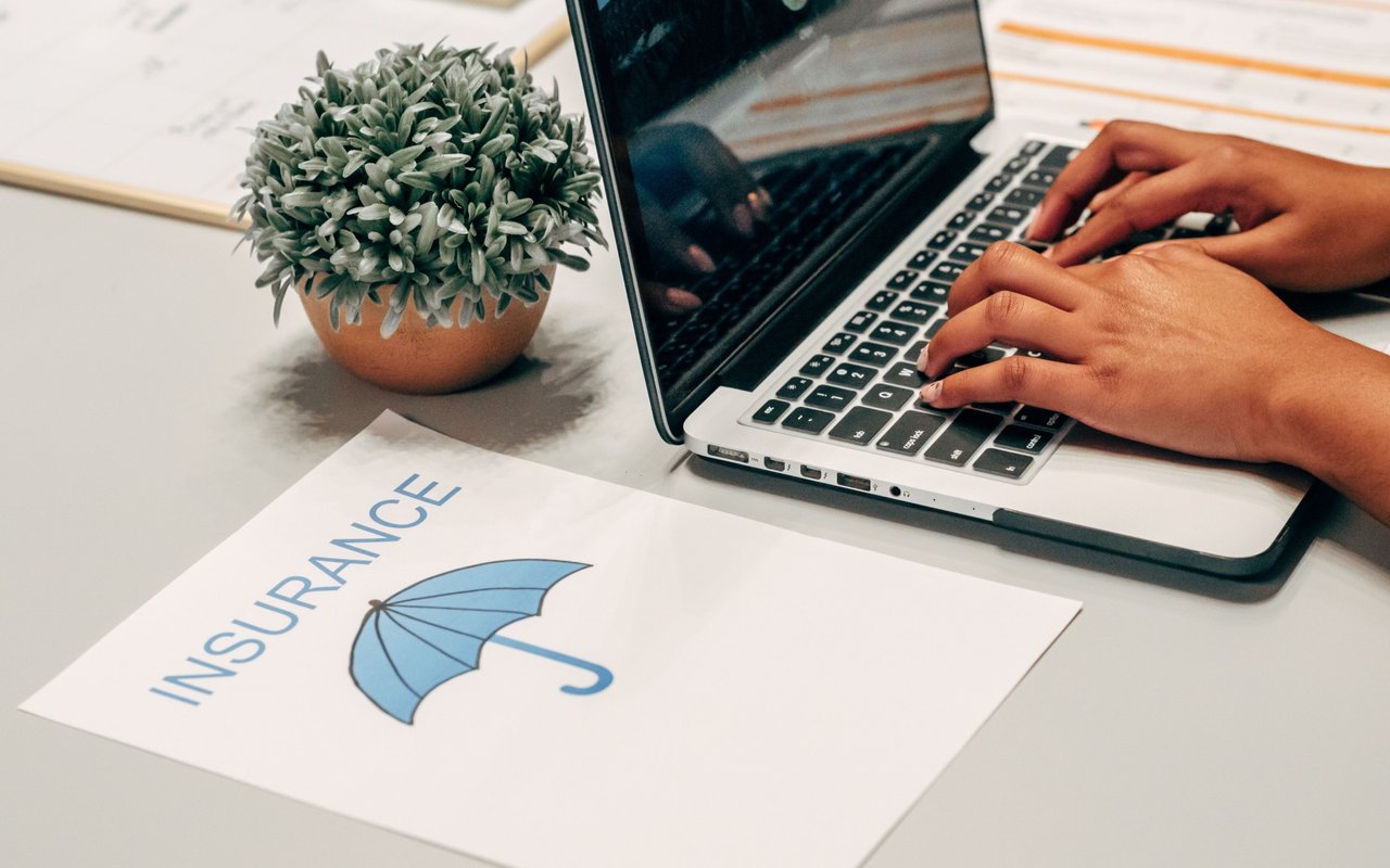 Hands type on a laptop beside an insurance umbrella illustration and a small potted plant.