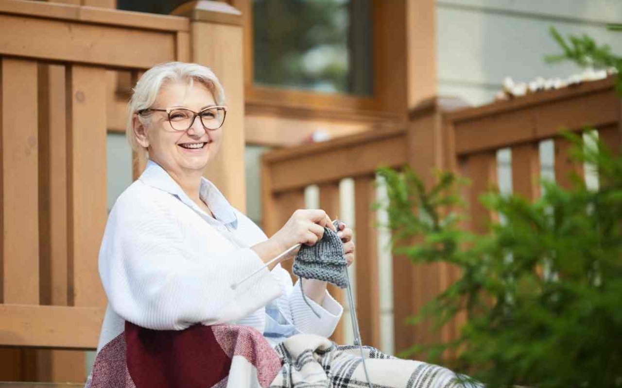 Older woman knitting on a wooden deck beside a house.