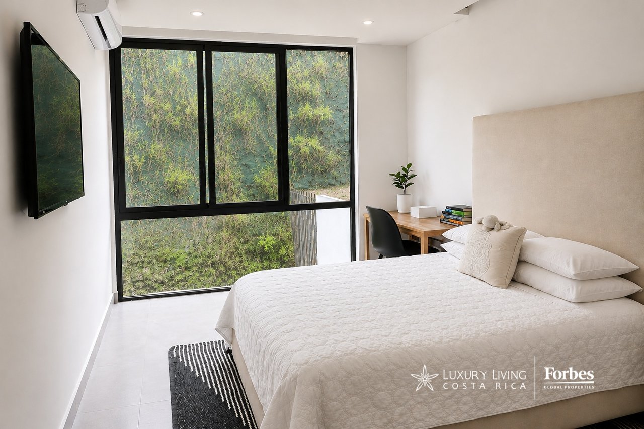 Contemporary bedroom with a white bed, beige headboard, and large windows overlooking greenery.