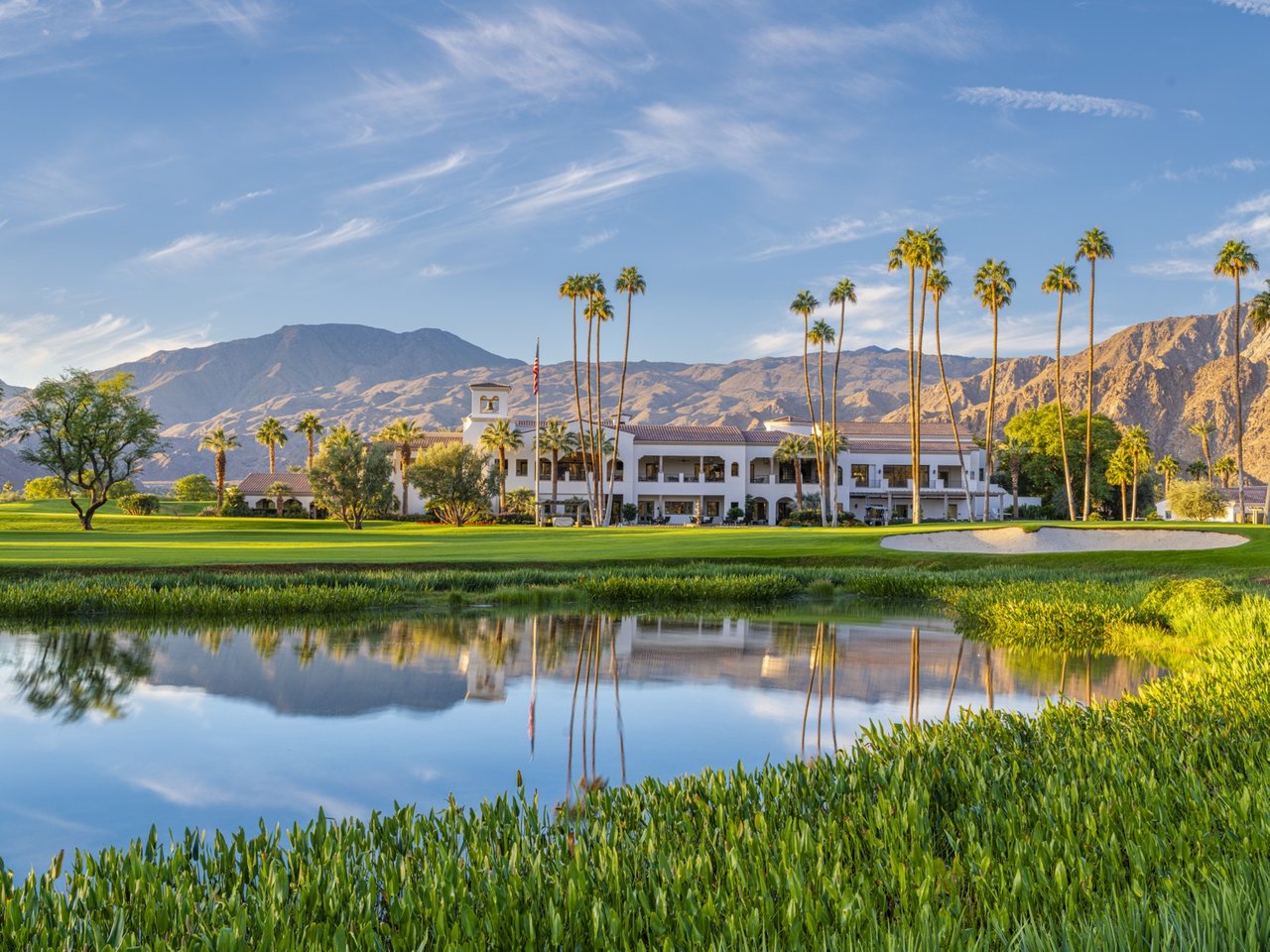 Golf clubhouse beside a reflective pond, framed by palm trees and desert mountains.