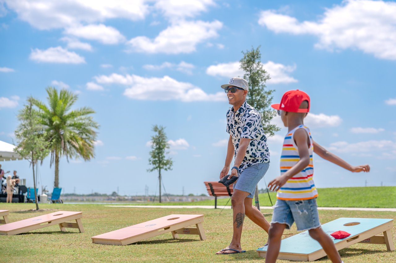 A man and boy play cornhole on a sunny lawn beside palm trees.