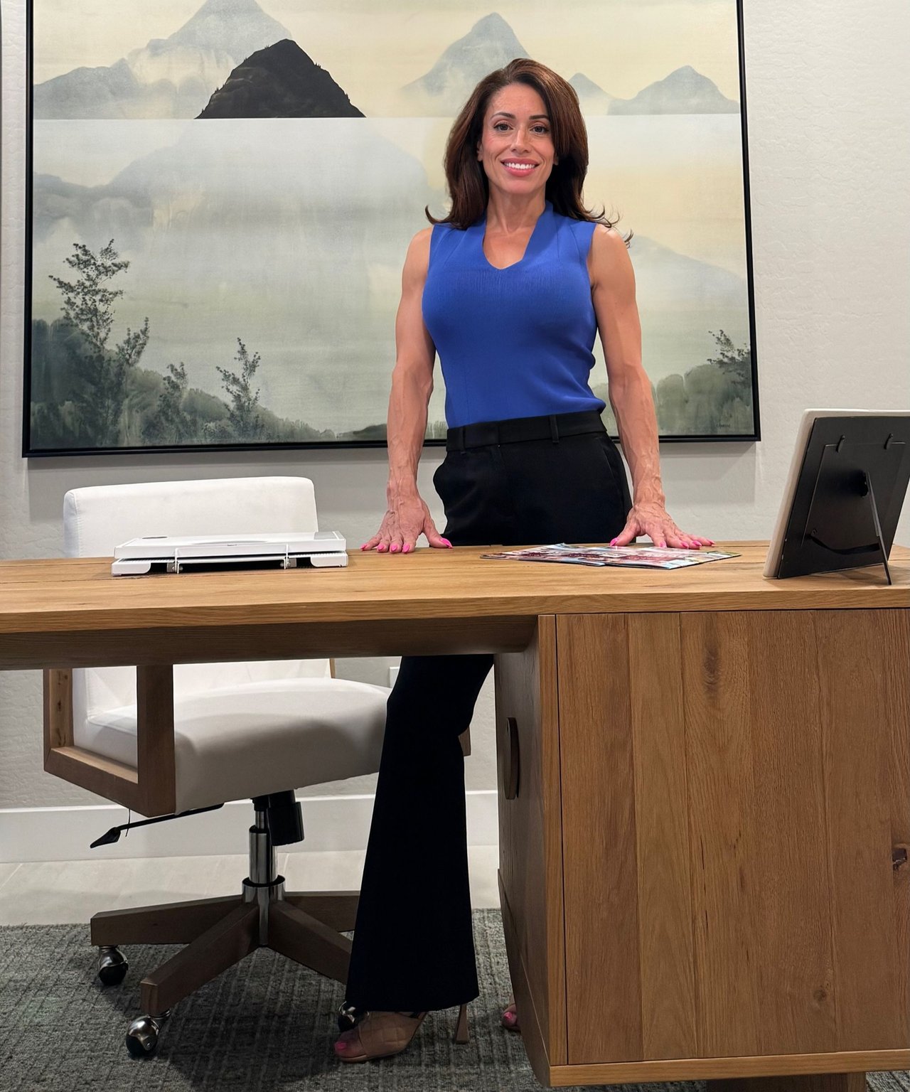 Gina Hall stands behind an office desk in a blue sleeveless top and black trousers.
