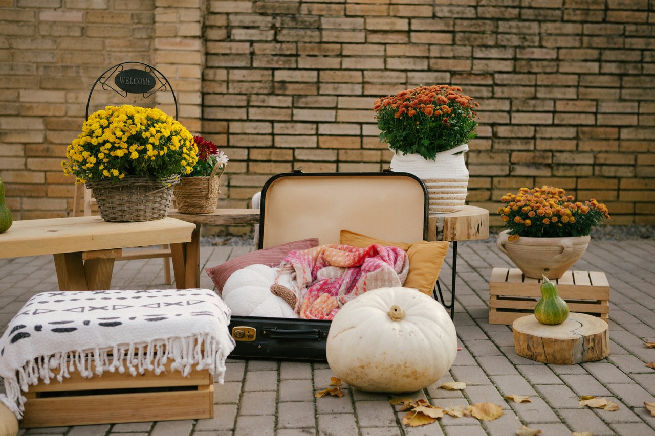 Open suitcase with colorful blankets, surrounded by autumn flowers and pumpkins on a brick patio.