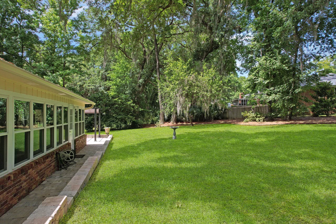 A lush green backyard with a lawn and large trees, bordered by a brick path and house on the left. The setting feels serene and inviting.