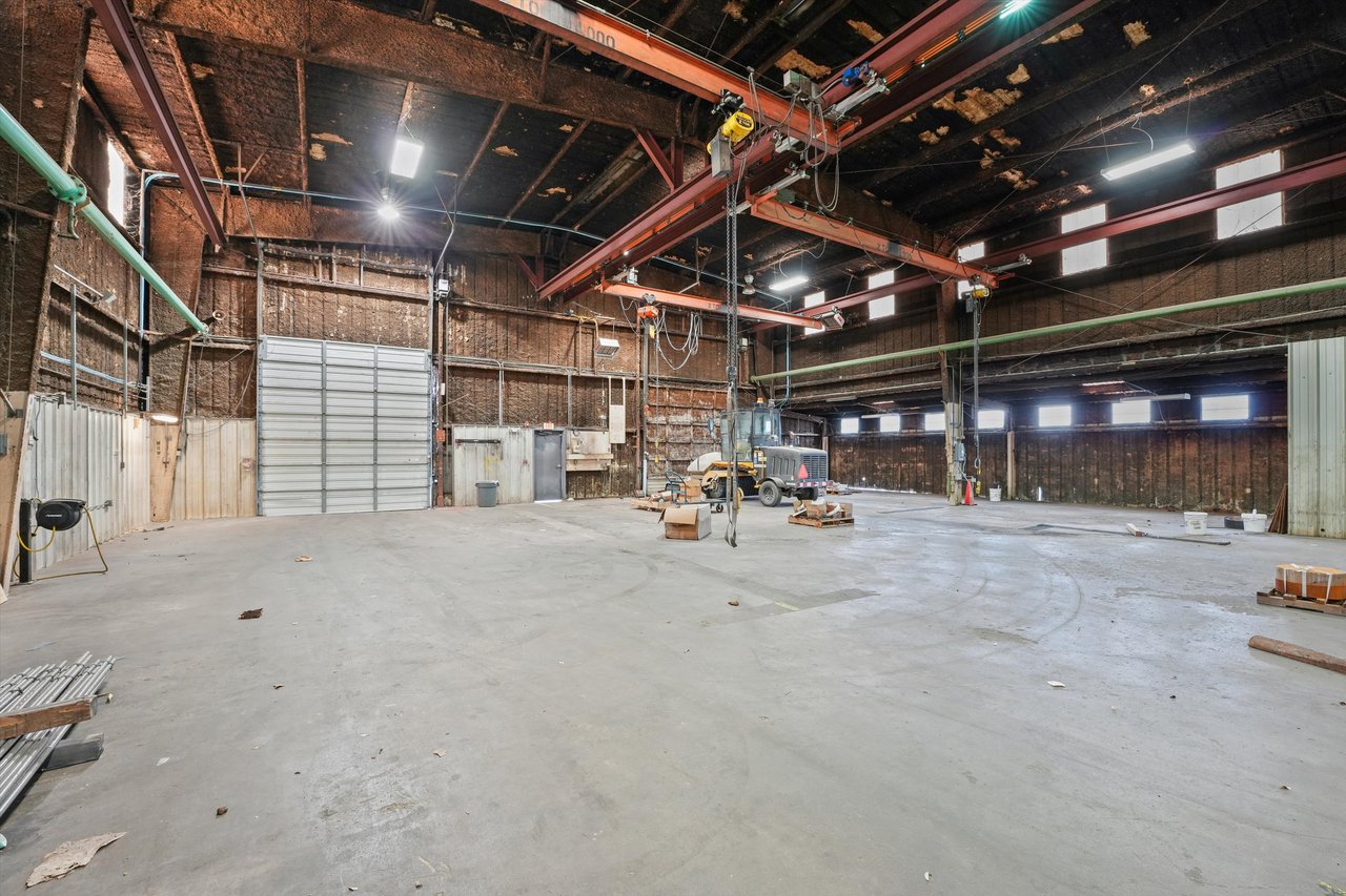 Industrial warehouse with exposed steel trusses, overhead lifting equipment, and a large roll-up door.