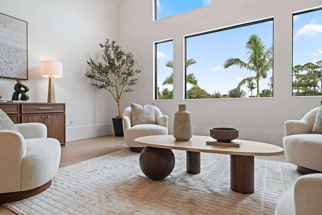 Furnished living room with tall windows overlooking palm trees and sculptural wood furniture.