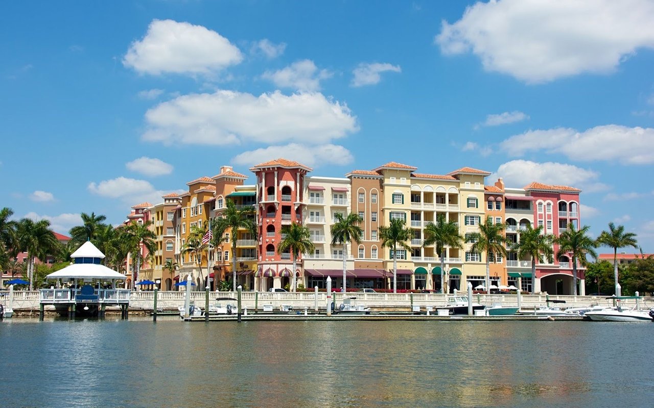Colorful waterfront buildings and palm trees line a marina with boats moored along the dock.