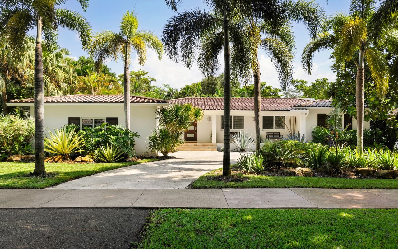 White, tile-roofed home with a paved driveway, palm trees, and tropical landscaping.