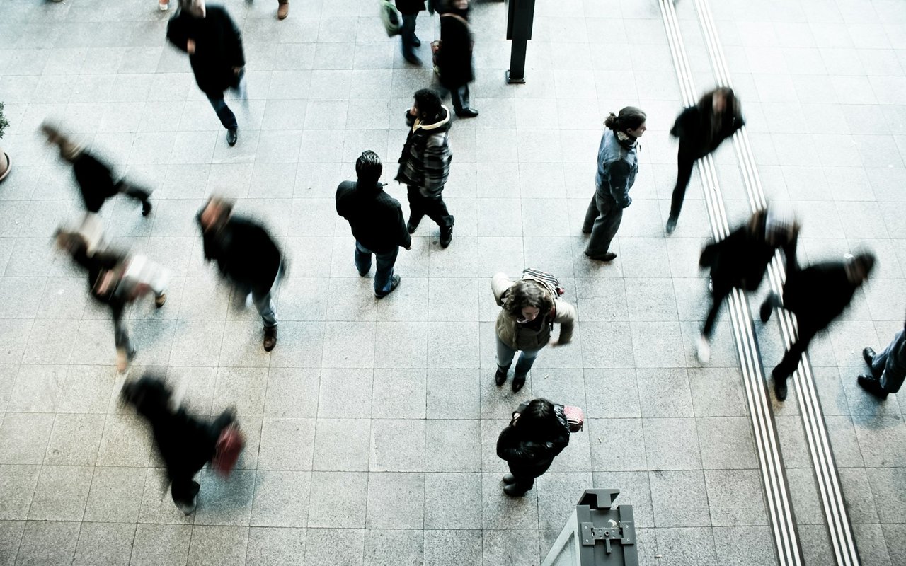 People cross a tiled indoor concourse, viewed from above, with several commuters blurred in motion.