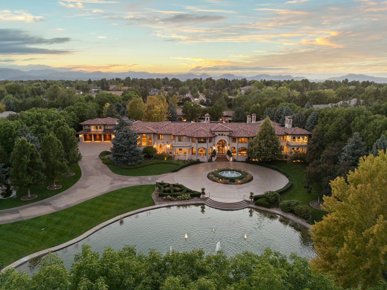 Aerial view of a sprawling estate with a circular driveway, fountain, pond, and distant mountains at sunset.