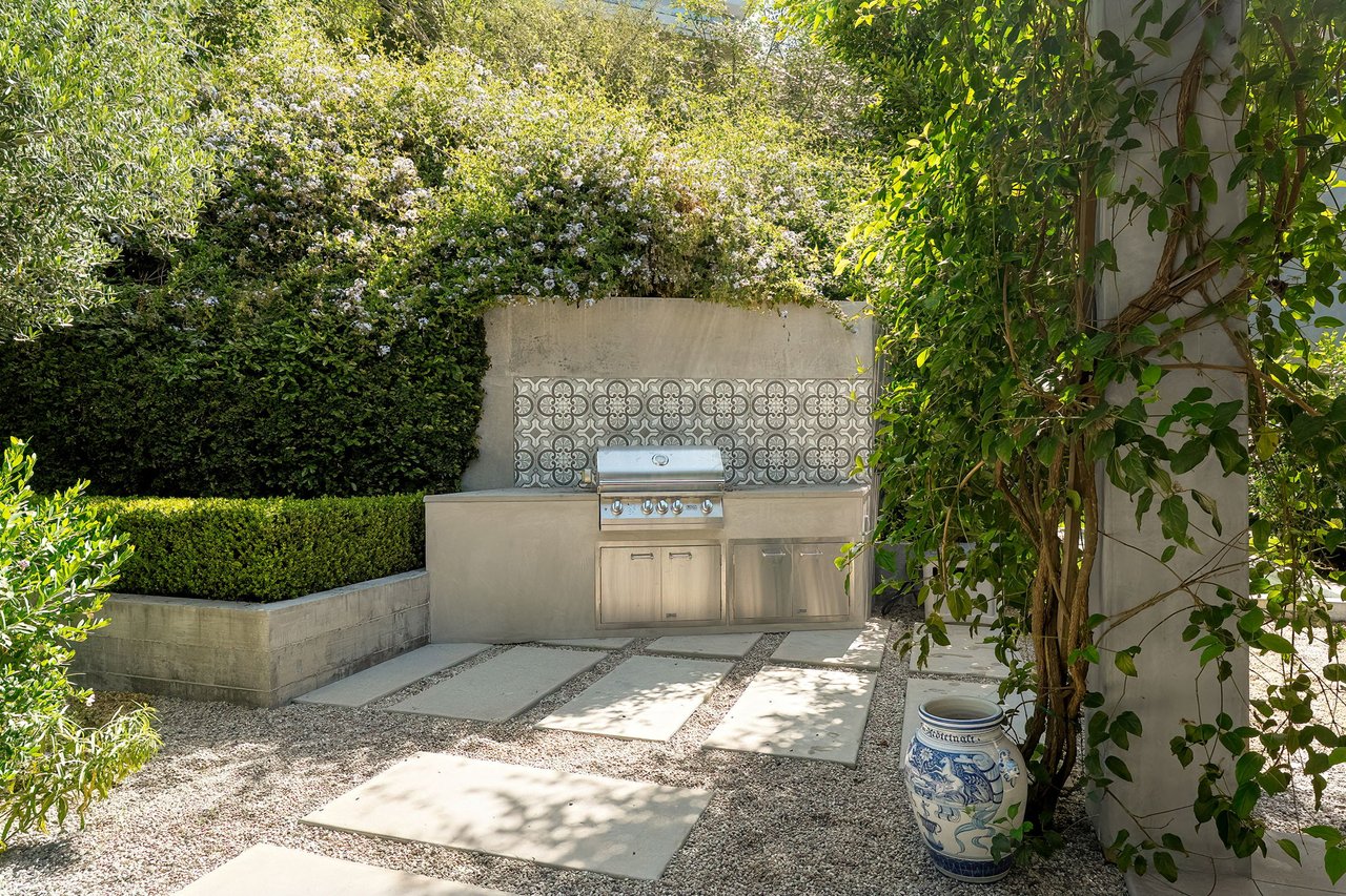 Outdoor kitchen with a stainless steel grill, patterned tile backsplash, and surrounding hedges.