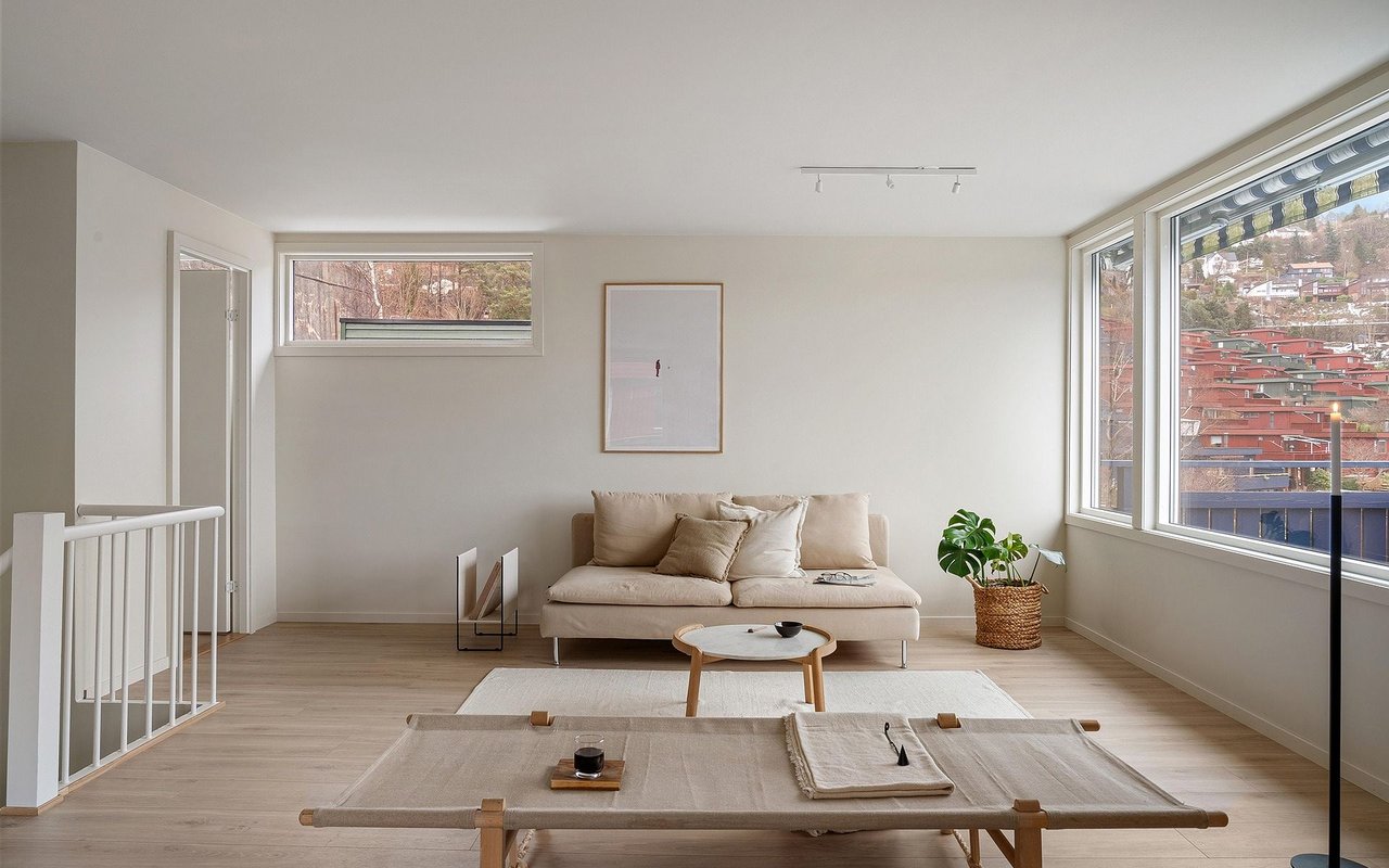 Living room with a beige sofa, pale wood floors, and large windows overlooking a hillside neighborhood.