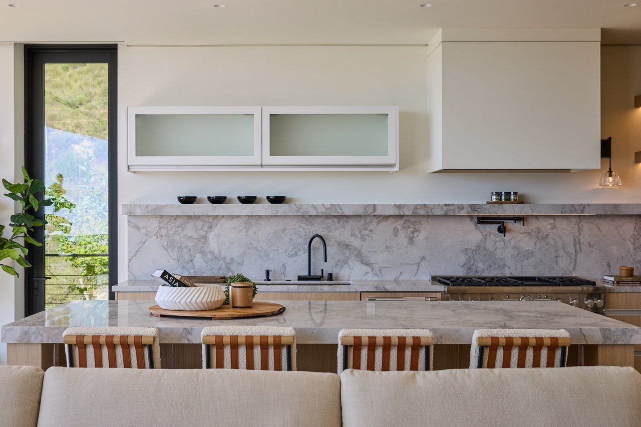 Modern kitchen with a marble island, matching backsplash, white cabinetry, and glass door to greenery.