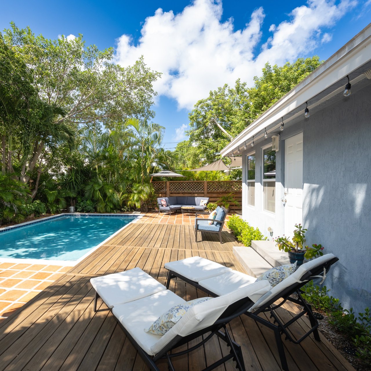 Backyard pool beside a wood deck with cushioned loungers and lush tropical landscaping.