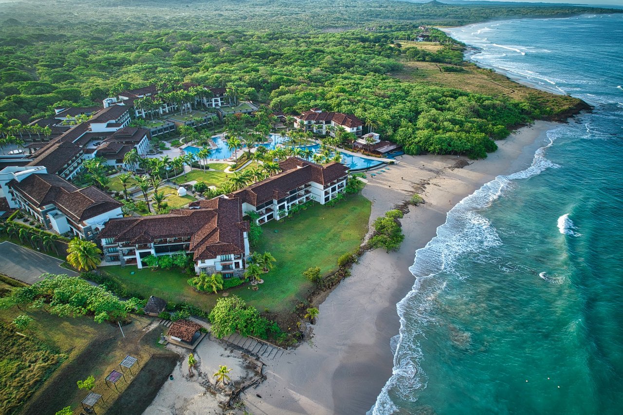 Aerial view of a beachfront resort with pools, tropical greenery, and a sandy Pacific shoreline.