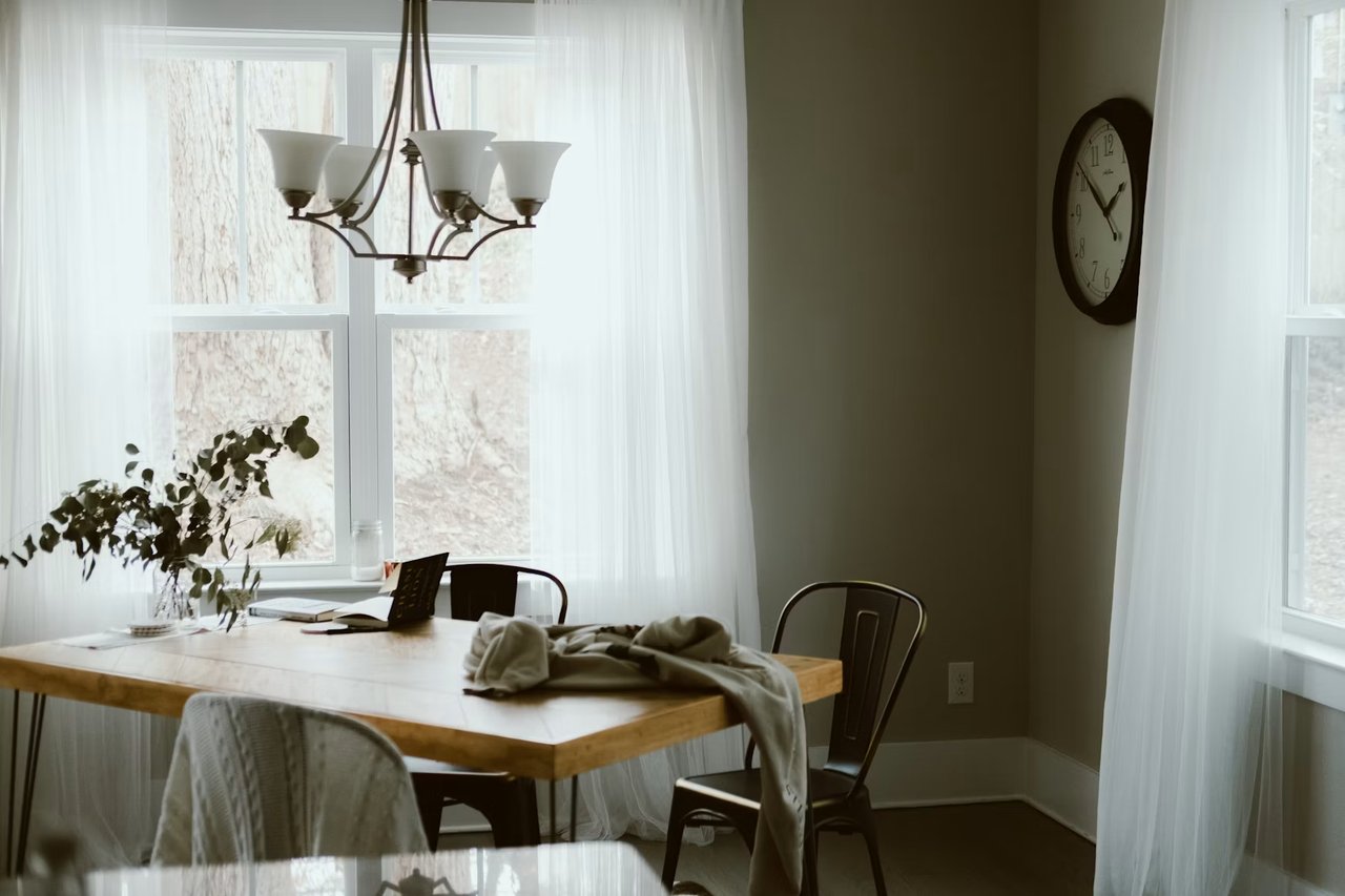 Dining room with a light wood table, sheer-curtained windows, and a chandelier.