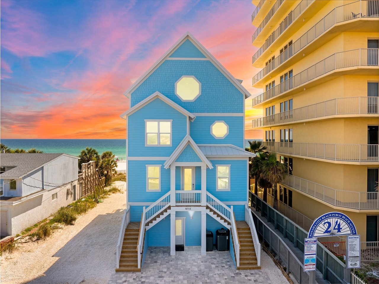 A stunning three-story light blue beach house with white trim located right next to Panama City Beach Public Beach Access 24 at sunset.