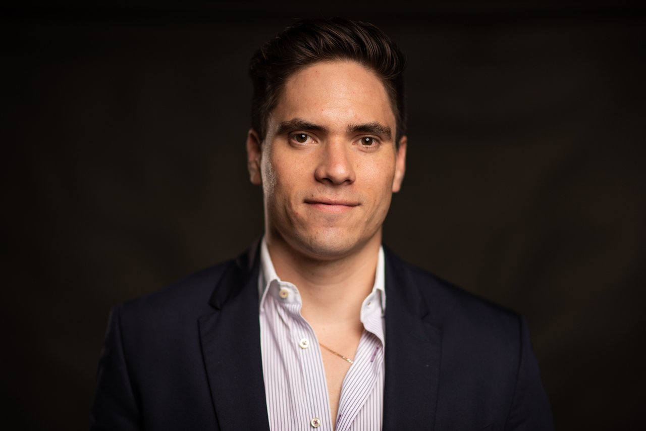 Man in a dark blazer and open-collar striped shirt against a dark background.