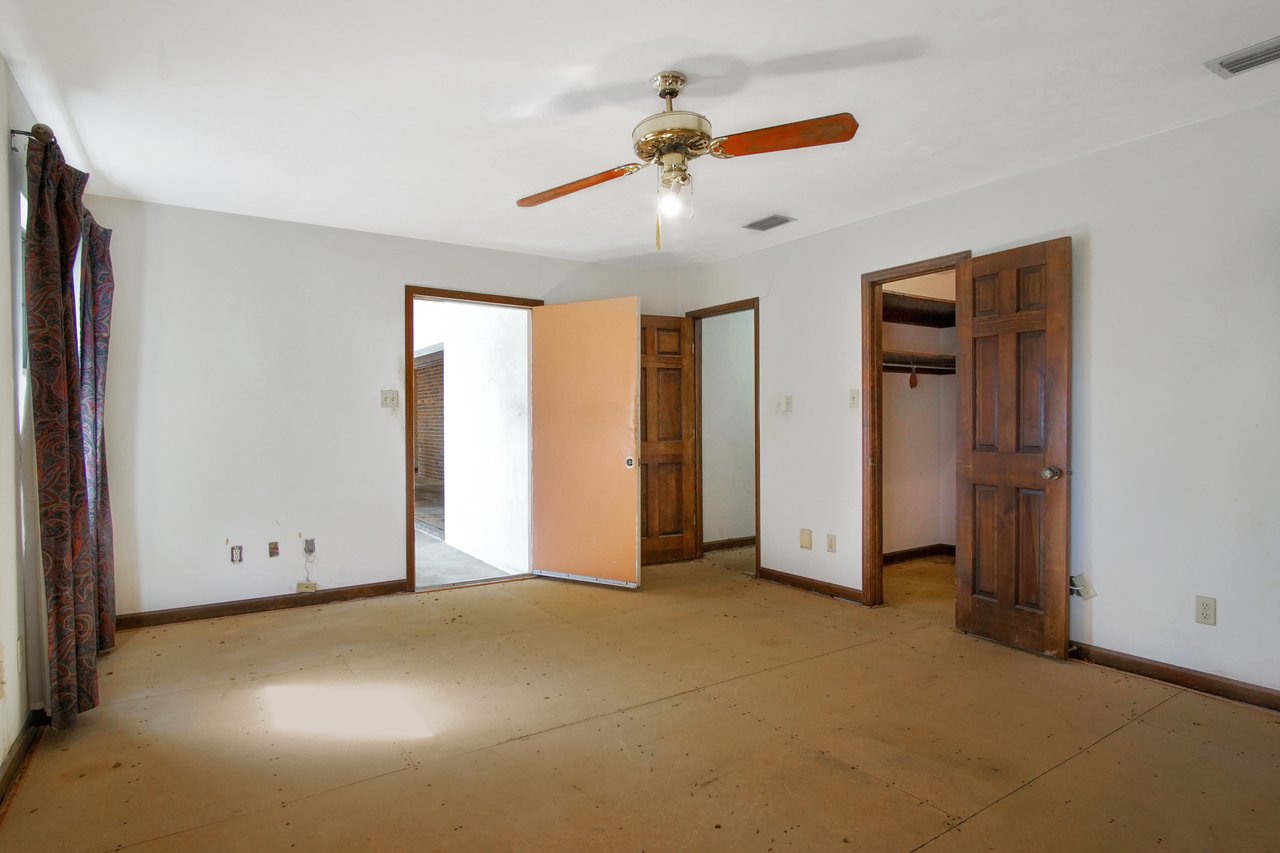 Empty room with white walls, an orange door partially open, exposing a hallway with brick walls. Wooden doors lead to a closet. A ceiling fan hangs above.